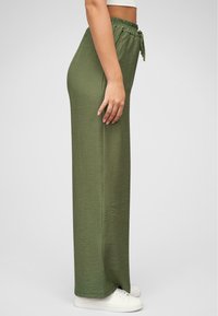 Pantalones verdes de pierna ancha hechos de una tela suave, con una cintura elástica y cordón. Se llevan con zapatillas blancas, se muestra el perfil lateral.