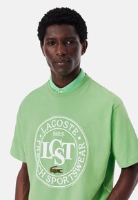 Camiseta de algodón verde con un logo redondo en blanco, que presenta "LACOSTE PARIS" y un cocodrilo bordado. Mangas cortas y un corte relajado.