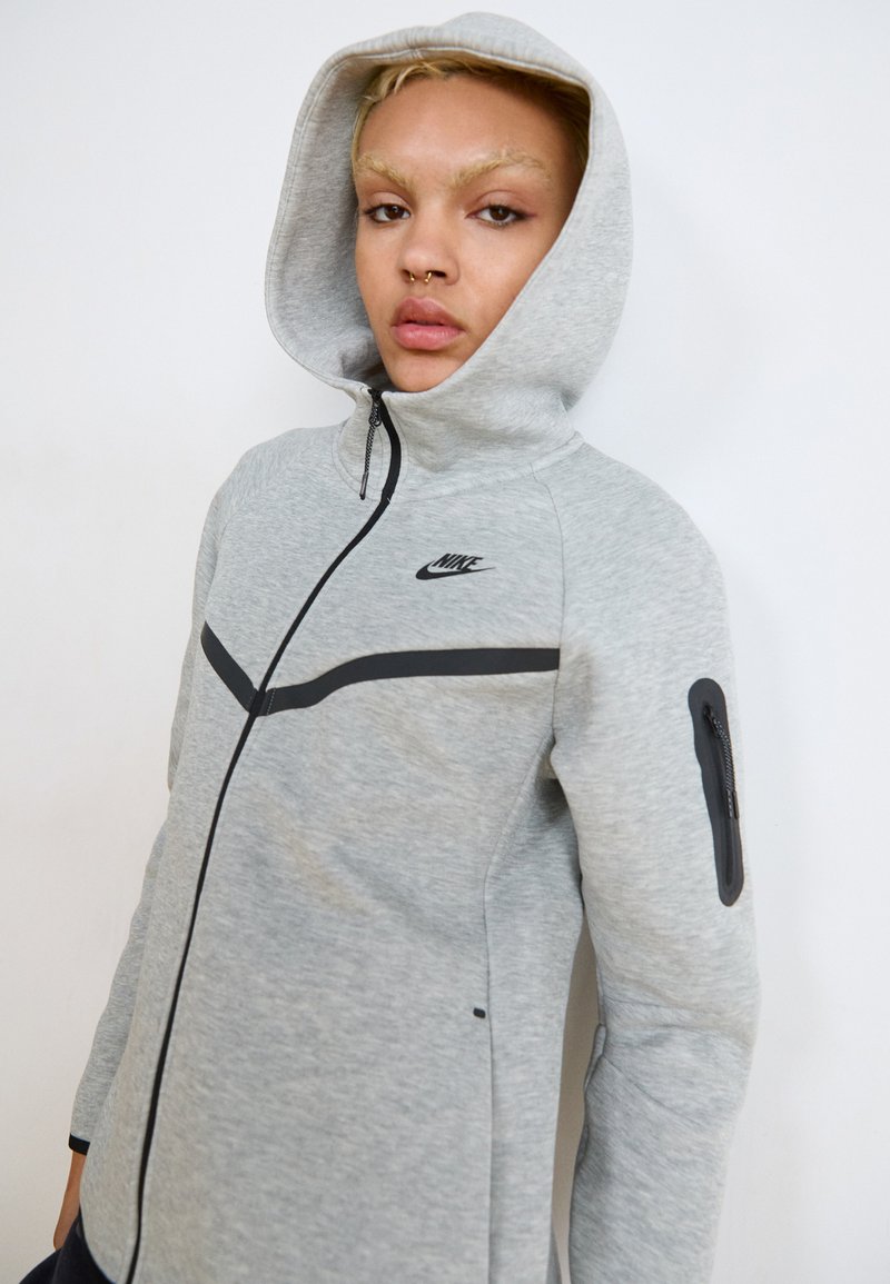 Grå zip-up hoodie lavet af blødt materiale, med hætte, sorte accenter og en lynlåslomme på venstre ærme. Nike-logoet er vist.