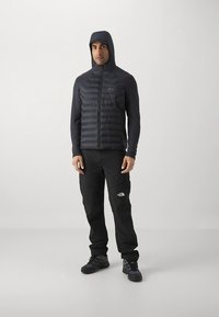 Svart isolert jakke med hette, med quiltet design. Kombinert med svarte bukser og svarte sko, egnet for utendørsaktiviteter.