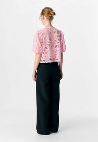Femme avec les cheveux en chignon portant un chemisier en dentelle florale rose transparent avec des manches bouffantes et un pantalon large taille haute noir, vue de dos.