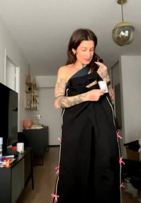 Kvinde med tatoveringer iført en sort beklædning med lyserøde sløjfer, som justerer en mærkat i en moderne stue med hylder og en pendellampe.