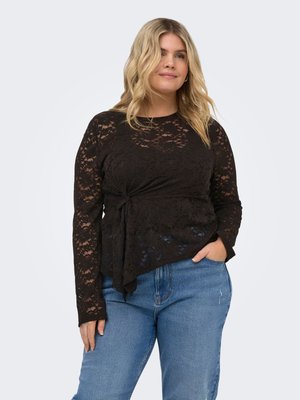 Femme aux cheveux blonds ondulés porte un top en dentelle noire à manches longues avec un nœud sur le devant et un jean en denim bleu, debout avec une main dans la poche.