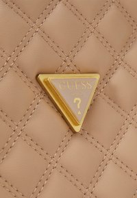 Sac à main matelassé marron clair avec un logo triangulaire doré bien visible portant l'inscription "GUESS" et un point d'interrogation. Surface texturée avec des détails de couture.