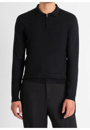 Uomo che indossa un maglione polo nero aderente a maniche lunghe con cerniera frontale e pantaloni neri, in piedi su sfondo bianco.