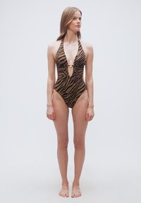 Γυναίκα όρθια, ξυπόλητη, φοράει καφέ και μαύρο ολόσωμο μαγιό με λωρίδες ζέβρας και λαιμόκοψη halter, με λεπτομέρεια μπροστά σε μορφή κρίκου, κοιτάζει ελαφρώς πλάγια.