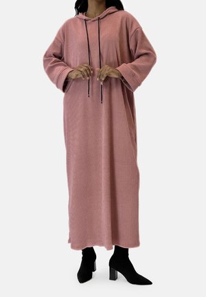 Femme portant une robe ample longue à capuche côtelée rose poudré avec des manches larges et des bottines noires à talons mi-mollet sur un fond uni.
