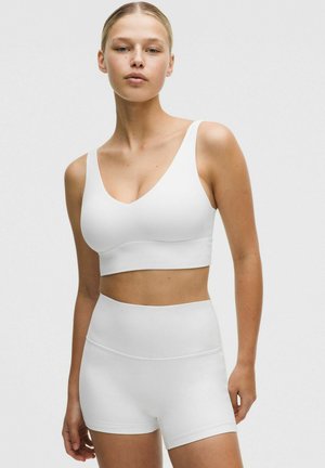 Kvinde iført en hvid sports-bh og matchende cykelshorts med høj talje, stående mod en ensartet lys baggrund.