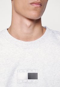 Sudadera gris claro con cuello redondo. Presenta un parche de logo elevado en blanco y negro, con acentos de rayas texturizadas. Textura de tela suave.
