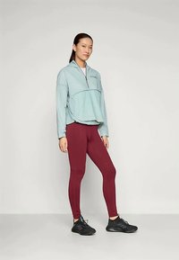 Jasnoniebieski rozpinany polar z kontrastującą ciemniejszą sekcją w talii, zestawiony z burgundowymi legginsami z przeszytymi liniami i czarnymi butami sportowymi.