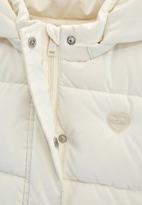 Cremfarbener, gesteppten Jackenbereich mit Reißverschluss, Druckknöpfen und herzförmigem Guess-Logo auf strukturiertem Stoff.