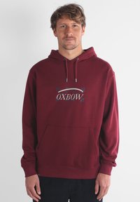 Haut à capuche bordeaux en tissu doux, avec une poche avant, une capuche à cordon et un logo avec des éléments décoratifs en bleu.