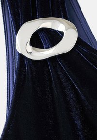 Tejido de terciopelo azul marino con una textura suave, diseño acanalado asegurado por una pieza de metal ovalada plateada brillante en el centro.