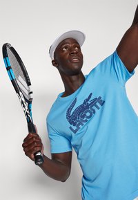 Lyseblå t-shirt med mørkeblåt Lacoste krokodillelogo, bæret af en person med en tennisracket. Tilbehøret inkluderer en hvid visir.