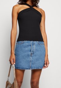 Mujer vistiendo un top halter negro y una minifalda de mezclilla azul, sosteniendo un bolso marrón texturizado a su lado.