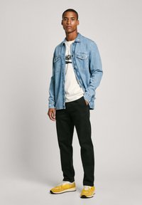 Denim košulja svijetloplave boje s dva džepa na prsima, bijela majica s crnom grafikom, crne hlače i žute tenisice s crnim detaljima.