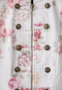 Veste blanche à motifs floraux roses et verts, fermeture éclair centrale et six boutons décoratifs en laiton ancien en deux rangées verticales.