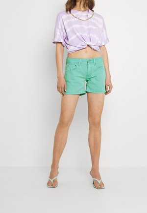 Světle fialové tričko s batikovaným vzorem a ohrnutými rukávy, kombinované se zelenými džínovými šortkami. Má klasické kapsy a volný střih. Mátově zelené sandály.