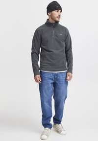 Grå fleece genser med kvart glidelås, brukt over en hvit skjorte, sammen med blå jeans og hvite joggesko, komplementert med en svart strikket lue.