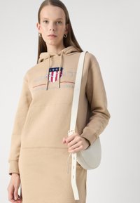 Beige hoodie with a front pocket, featuring a logo in red and blue. The model holds a light gray crossbody bag with a strap.