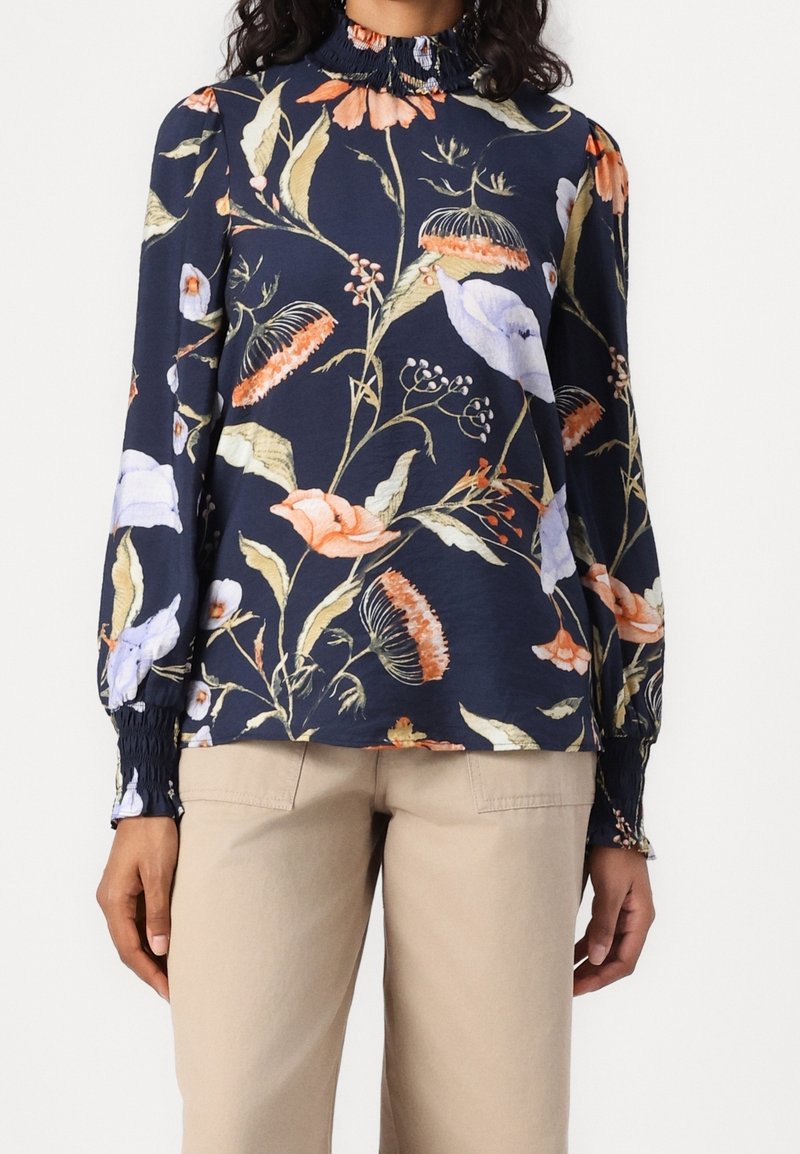 Femme portant un chemisier bleu marine à manches longues avec un col montant à volants et un grand imprimé floral, associé à un pantalon beige.