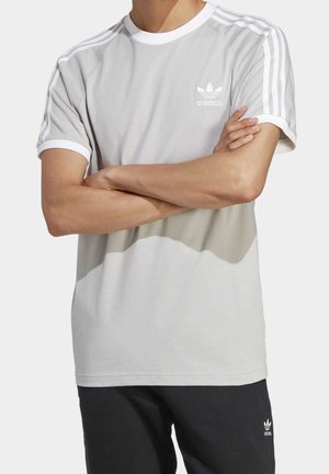 Personne portant un t-shirt Adidas gris clair avec des rayures blanches sur les manches et un pantalon Adidas noir, les bras croisés sur la poitrine.