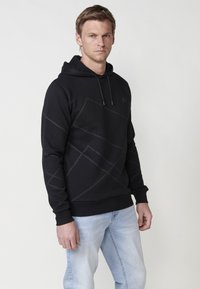 Hanorac negru cu modele geometrice gri, glugă cu șnur și manșete ribbate; purtat cu blugi de un albastru deschis. Textură a materialului neted.