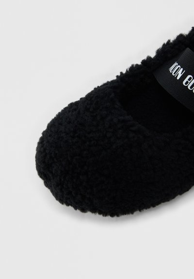 Chausson noir duveteux à bout arrondi avec une étiquette intérieure visible sur un fond blanc uni.