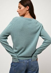 Frau mit langen braunen Haaren, die einen hellblauen Pullover und blaue Jeans trägt, steht mit den Händen in den hinteren Taschen und blickt zur Seite.