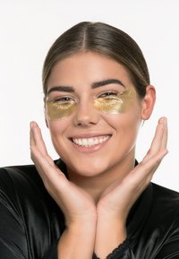 Gullunder-eye patches på en persons ansikt, med lys hudfarge og svart satin morgenkåpe, smilende med hendene plassert under haken.