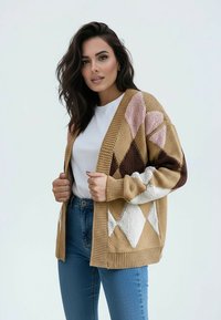 Kardigan z dzianiny z wzorem diamentowym w kolorach beżowym, brązowym, różowym i białym. Otwarty przód, wykończony ściągaczami na mankietach i dole. Połączony z białą koszulą i niebieskimi dżinsami.