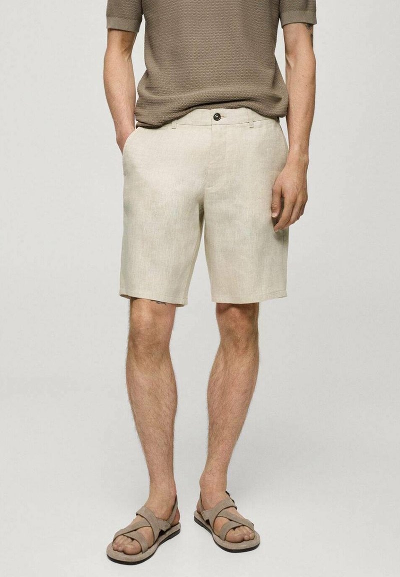 Homme portant un short beige mi-cuisses, une chemise marron texturée à manches courtes, et des sandales beige, debout les mains dans les poches.