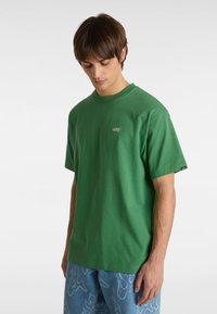 T-shirt in cotone verde con collo rotondo e maniche corte. Presenta un piccolo logo ricamato Vans sul petto. Abbinato a jeans blu con motivo.