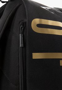 Rucsac negru cu o textură netedă, un logo auriu proeminent și un fermoar negru cu un tab de tragere texturat. Accente din plasă sunt vizibile.