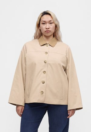 Jeune personne aux longs cheveux portant une veste beige boutonnée avec col et un jean bleu, debout devant un fond blanc uni.
