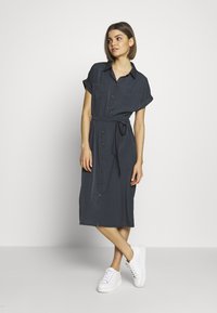 Abito camicia grigio scuro con maniche corte, chiusura a bottoni sul davanti, cintura da annodare in vita e due tasche frontali; indossato con scarpe da ginnastica bianche.