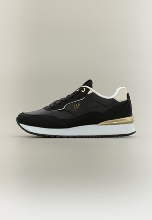 Zwart-witte sneaker met gouden accenten en "GAP"-logo aan de zijkant, laag uitgesneden ontwerp en dikke witte zool met zwart profiel.