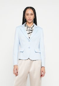 Blazer azul claro de diseño entallado, cierre de dos botones y bolsillos con solapa. Se lleva sobre una camisa con patrón en blanco y negro.