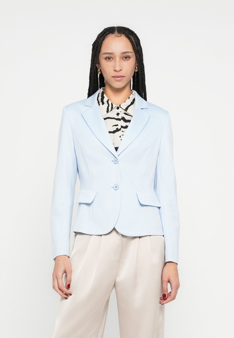 Blazer azul claro de diseño entallado, cierre de dos botones y bolsillos con solapa. Se lleva sobre una camisa con patrón en blanco y negro.