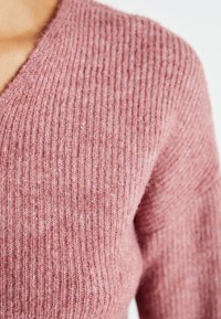 Weicher, gerippter Strickpullover in staubrosa Farbe mit einem V-Ausschnitt-Design, das einen strukturierten Stoff für zusätzliche Wärme und Detail zeigt.