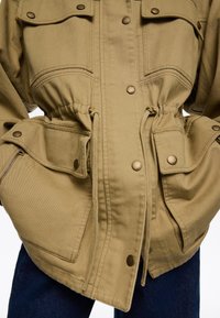 Chaqueta caqui de corte relajado, con dos bolsillos delanteros, botones de presión metálicos, cintura ajustable con cordón y una textura de tela resistente.
