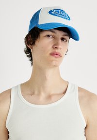 Gorra trucker azul y blanca con un parche de logo, paneles laterales de malla y visera curva. Llevada con una camiseta de tirantes blanca acanalada.