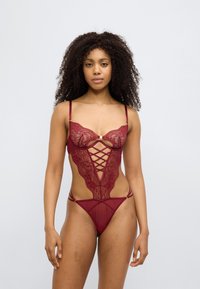 Κορμάκι από μπορντό δαντέλα με βαθιά λαιμόκοψη, λεπτομέρειες με σταυρωτούς ιμάντες και διάφανα πλαϊνά πάνελ. Ρυθμιζόμενοι ιμάντες για καλύτερη εφαρμογή.