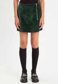 Green corduroy mini skirt with a side zipper, featuring a ribbed texture and a fitted design. Paired with knee-high black socks and loafers.