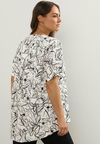 Donna che indossa una blusa bianca ampia con stampa floreale astratta nera, in piedi di lato contro un muro chiaro e semplice.