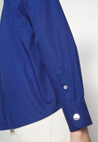 Detalle de una camisa de manga larga azul con puños puntiagudos con botones, textura suave y un diseño de bajo ligeramente curvado.
