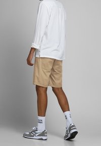 Pantalones cortos beige con bolsillos, combinados con una camisa blanca de manga larga. Zapatillas deportivas grises y blancas con calcetines de colores, de pie sobre un fondo claro.