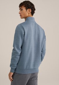 Ljusblå sweatshirt med hög krage, strukturerad tyg och ribbade ärmslut och nederkant. Modell sedd bakifrån, iförd grå byxor.