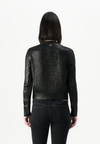 Femme avec des cheveux raides et foncés portant un pull en tricot noir brillant et un jean noir, se tenant de dos face à l'appareil photo sur fond blanc.