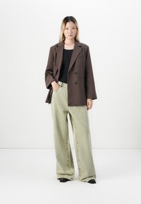 Jeune femme portant un blazer brun surdimensionné, un haut noir, un jean large vert clair et des chaussures noires à bouts pointus, se tenant contre un mur blanc.